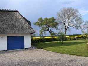 Une maison avec un toit de chaume et une porte de garage bleue, entourée d’une pelouse bien entretenue et d’un champ de fleurs jaunes.