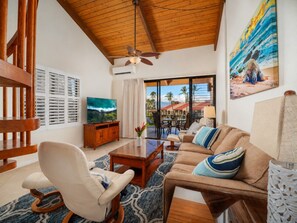 A living room with a wooden ceiling, a television, a sofa, a coffee table, and a view of the outdoors. - Walk to Beach! Pool, Gym, Pickleball & More (Kihei)