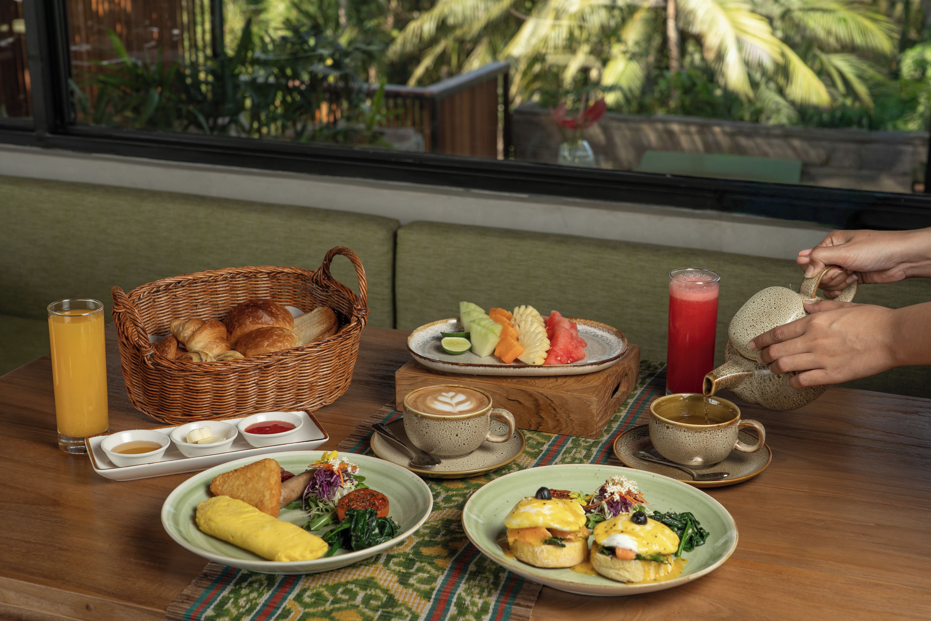 A breakfast setting with a wicker basket, croissants, fruit, coffee, and a person pouring tea.