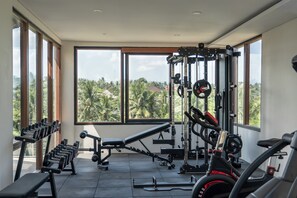 A gym with exercise equipment, large windows, and a view of palm trees. - Premadewi Resort A Pramana Experience (Ubud)