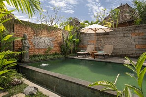 A swimming pool with a stone wall, a statue, and two lounge chairs with an umbrella. - Premadewi Resort A Pramana Experience (Ubud)