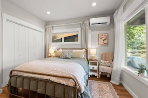 A bedroom with a bed, bedside tables, a chair, an air conditioning unit, and a window with curtains. - Little Garden Bungalow (Spokane)