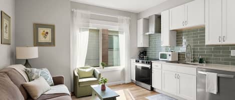 A compact living space with a sofa, a small table, a window with curtains, a kitchenette with a microwave, and a stove.