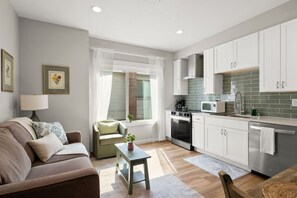 A compact living space with a sofa, a small table, a window with curtains, a kitchenette with a microwave, and a stove. - Little Garden Bungalow (Spokane)
