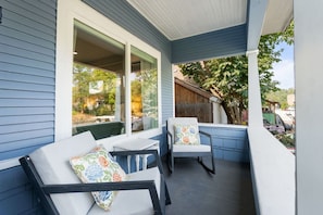 A covered porch with two chairs and a table, large windows, and a view of the backyard. - Little Garden Bungalow (Spokane)