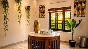 A room with a round wooden table, a vase with plants, a window with blinds, and a wall-mounted light fixture. - Maiibreeze Retreat (Sultan Bathery)