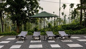 A set of outdoor lounge chairs with a green umbrella, arranged on a gravel surface with a backdrop of palm trees and dense foliage. - Maiibreeze Retreat (Sultan Bathery)