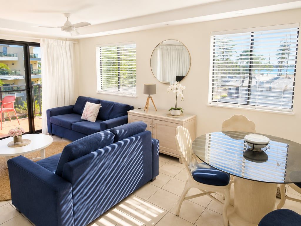 A modern living room with a blue sofa, a glass dining table, and a view of the outdoors through large windows.