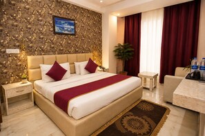 A hotel room with a bed, a nightstand, a desk, a chair, a potted plant, and a window with curtains. - Ektham Hotels (Haridwar)