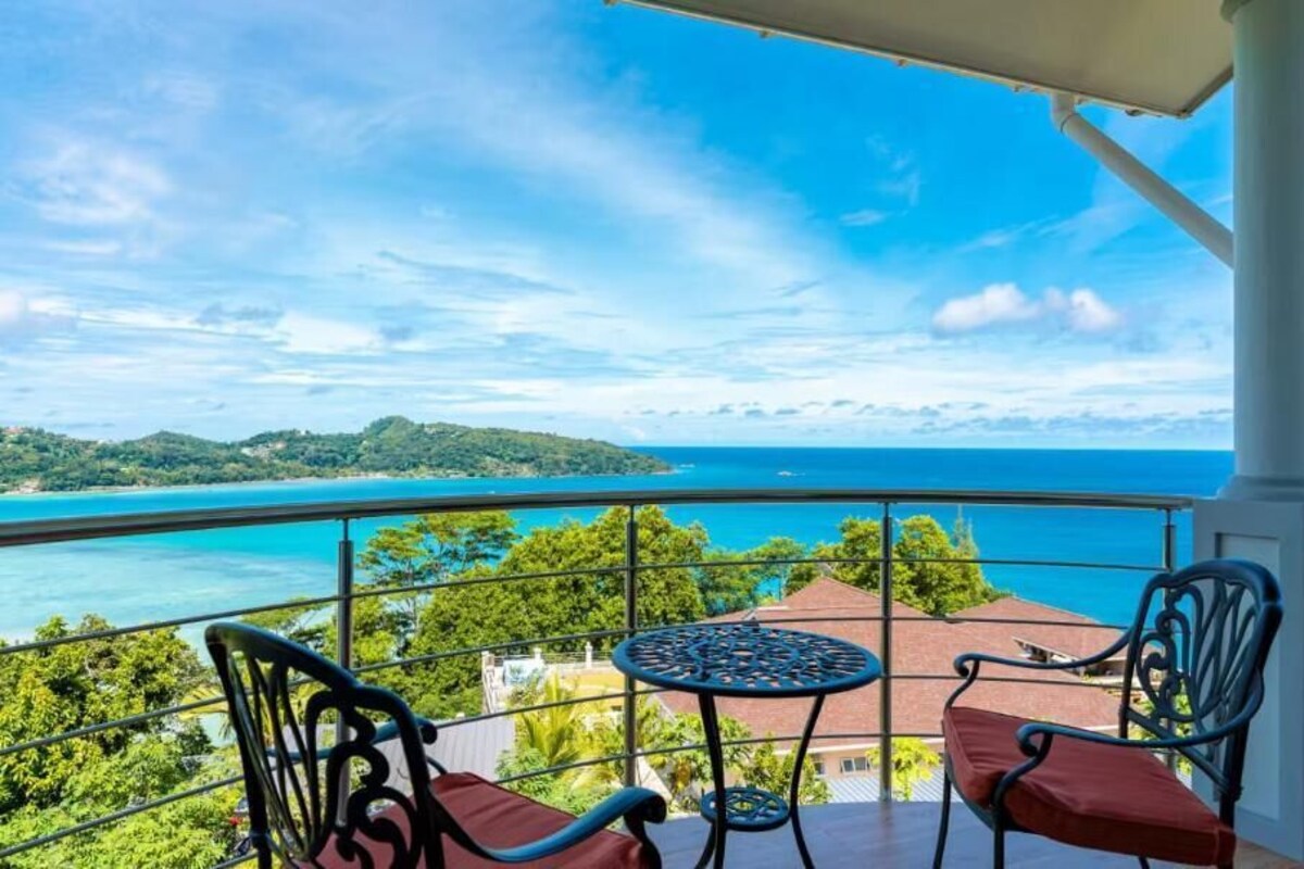 A balcony with a table and chairs, overlooking a coastal landscape with a clear view of the sea and distant land.
