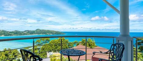 A balcony with a table and chairs, overlooking a coastal landscape with a clear view of the sea and distant land.