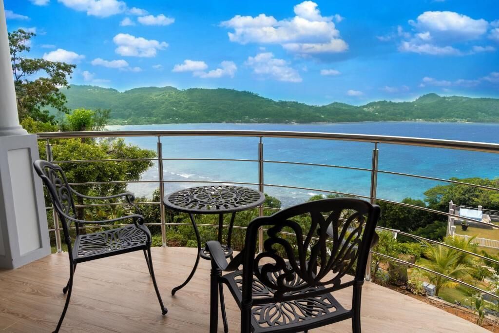 A balcony with a table and two chairs, overlooking a body of water and green hills.
