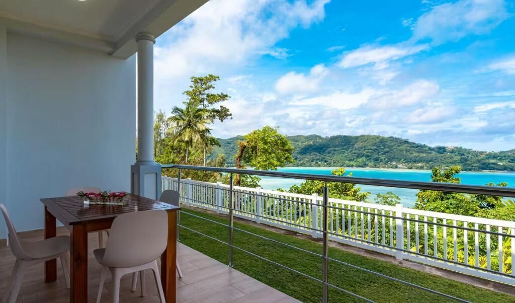 A balcony with a table and chairs, overlooking a turquoise sea and green hills.