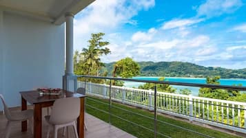 A balcony with a table and chairs, overlooking a turquoise sea and green hills.