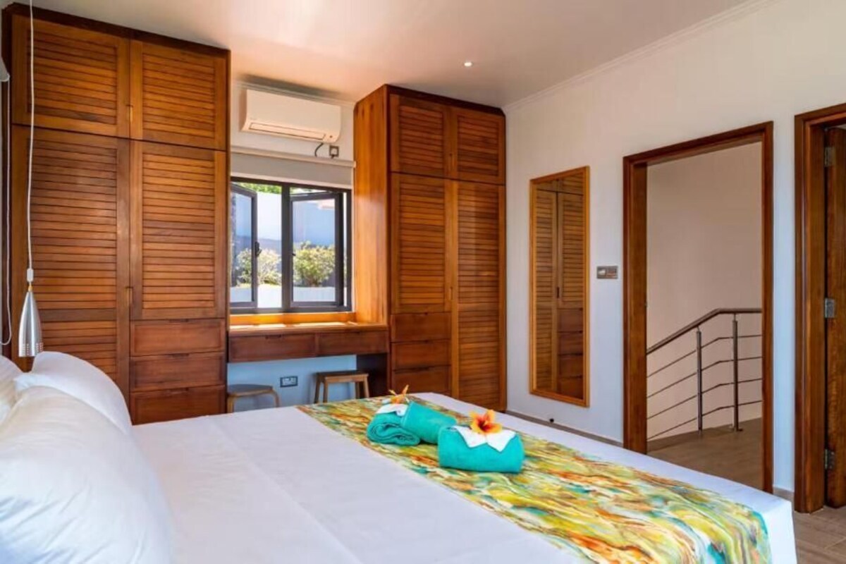 A hotel room with a bed, wooden wardrobe, a desk with a chair, a window, and a staircase.