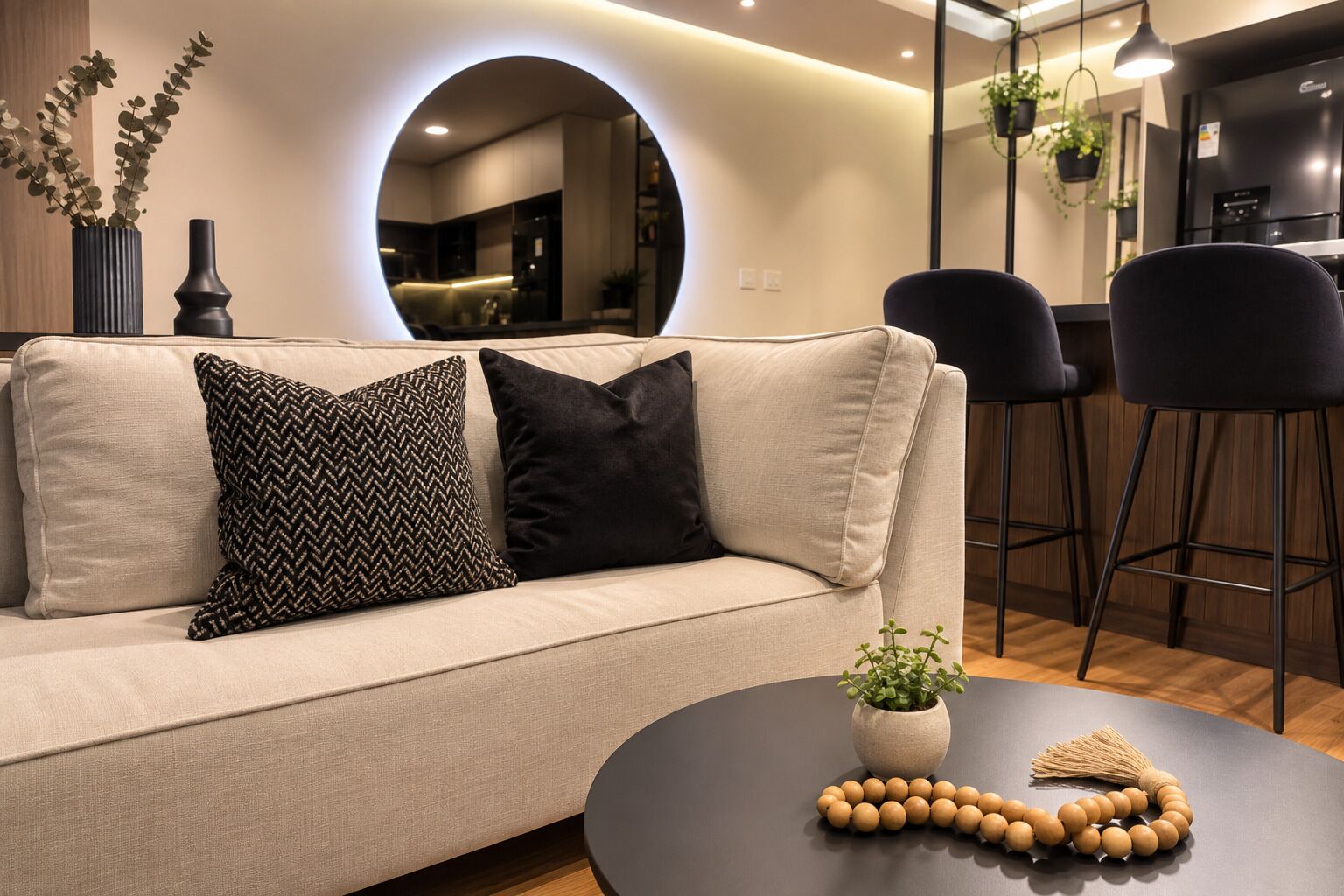 A modern living room with a sofa, two black pillows, a round table with a plant and a beaded object, and a kitchen area in the background.