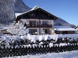 Um chalé de dois andares com uma varanda, rodeado por árvores e montanhas cobertas de neve.