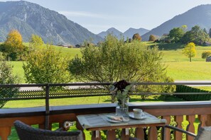 Uma varanda com mesa posta para refeição, com vista para uma paisagem verde com árvores e montanhas.