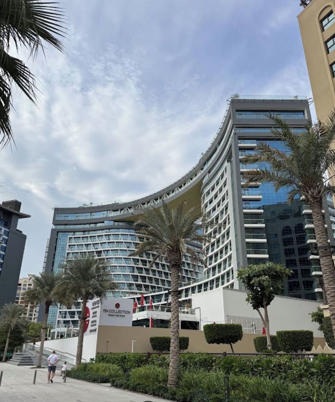 A modern hotel with a curved facade, palm trees, and a sign for 'The Collection Dubai Palm'.