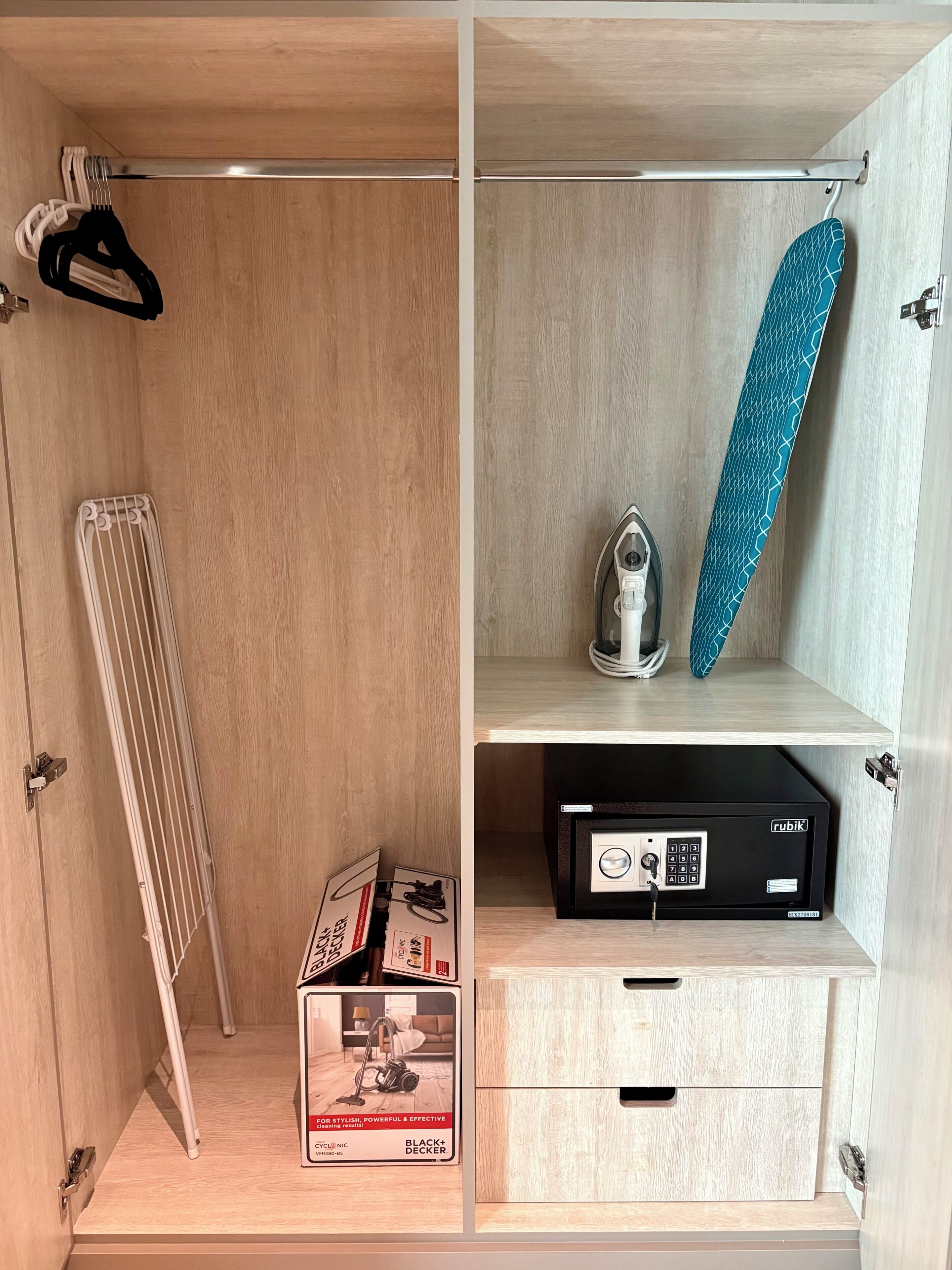 A room with a wooden floor, a radiator, a vacuum cleaner, a blue ironing board, and a black security safe.