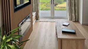 A modern living room with a flat-screen TV mounted on a wooden panel wall, a large sliding glass door leading to a scenic outdoor view, and a wooden coffee table with a notebook and remote control. - Barlocco -  a lodge that sleeps 2 guests  in 1 bedroom (Isles of Fleet Retreat, Gatehouse of Fleet)