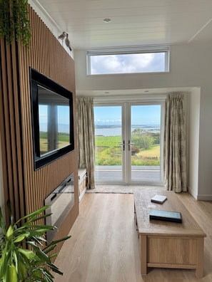 A modern living room with a flat-screen TV mounted on a wooden panel wall, a large sliding glass door leading to a scenic outdoor view, and a wooden coffee table with a notebook and remote control. - Barlocco -  a lodge that sleeps 2 guests  in 1 bedroom (Isles of Fleet Retreat, Gatehouse of Fleet)
