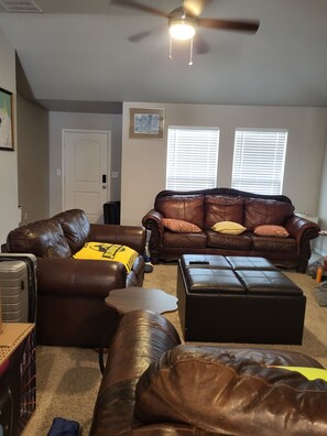 A living room with a brown leather sofa, a matching ottoman, and a ceiling fan. - EvidenceTouch Homes 03 (Odessa)