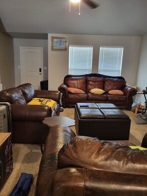 A living room with a brown leather couch, a matching ottoman, and a small table. There are two windows with blinds, a door, and a ceiling fan.