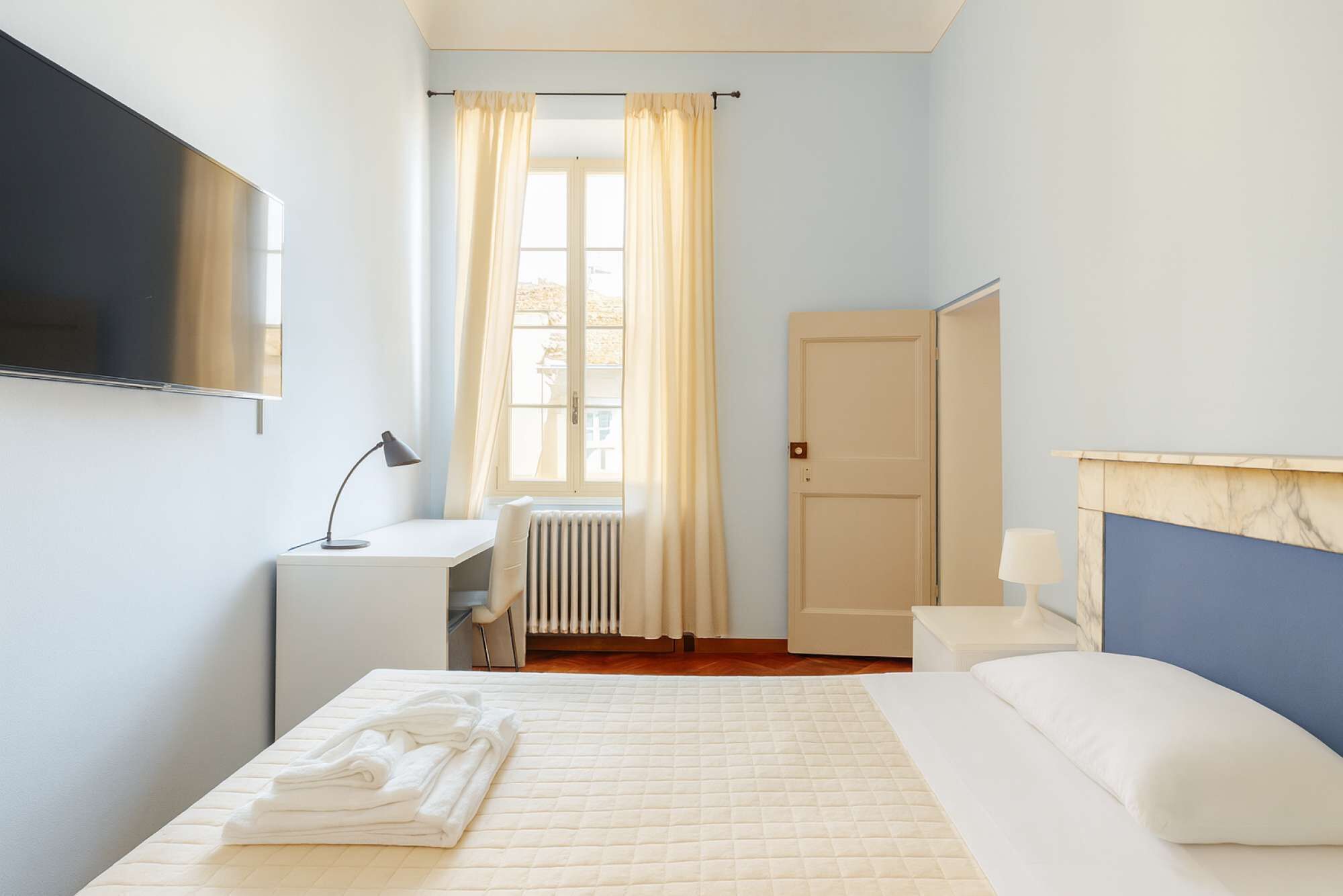 Ein Hotelzimmer mit Bett, Schreibtisch, Stuhl, Heizkörper, Wandfernseher und einer Tür.