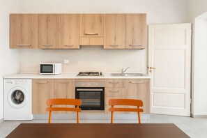 Une cuisine compacte avec des meubles en bois, un four à micro-ondes, une cuisinière, un évier et un lave-linge. On y trouve deux chaises orange et une table au centre.