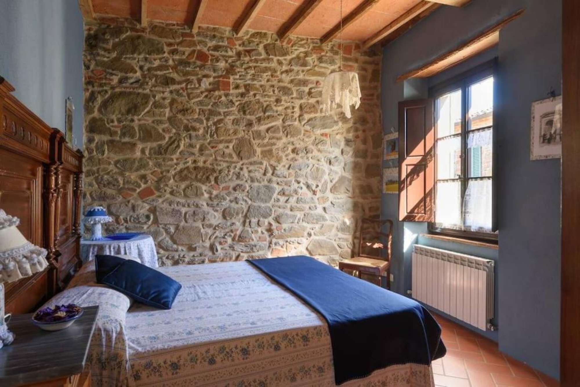 A bedroom with a stone wall, a bed with a blue bedspread, a wooden dresser, and a window with shutters.
