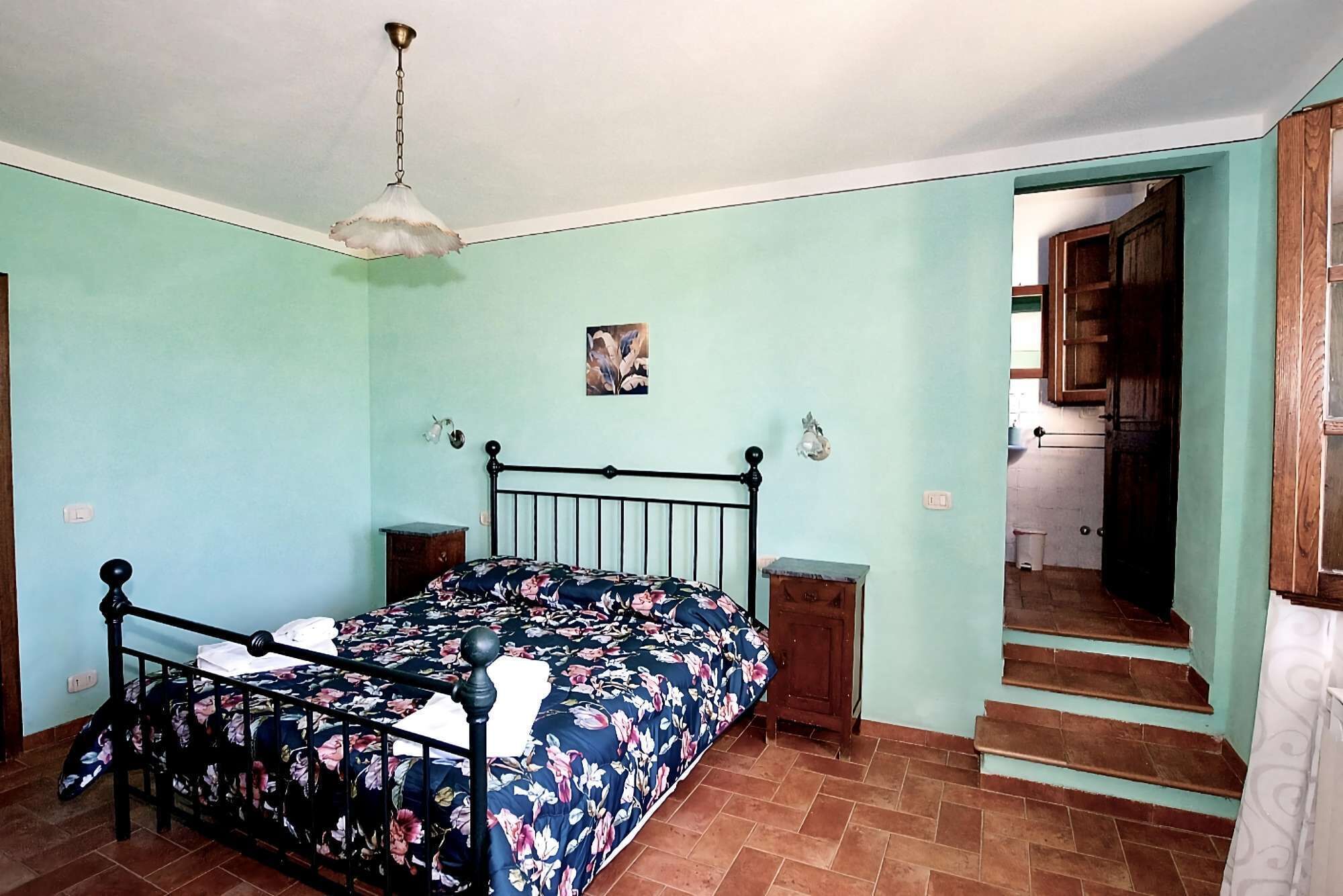 Un dormitori amb un llit, una tauleta de nit, una làmpada penjada a la paret, un quadre petit i una escala que porta a una altra habitació.