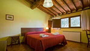A bedroom with a wooden headboard, a bed with red bedding, a bedside table, a lamp, a window with curtains, and a radiator.