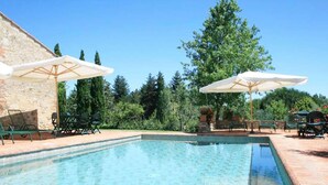 A swimming pool with lounge chairs and umbrellas, surrounded by greenery and a stone wall.