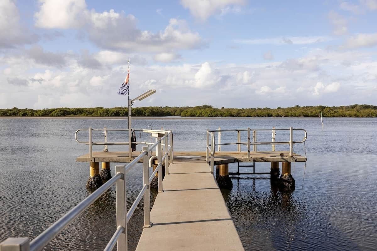 A concrete pier extending into calm water with railings, a flag, and a clear sky.