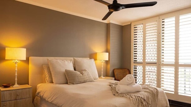 Quarto com cama, duas luminárias de cabeceira, ventilador de teto e janela com persianas.