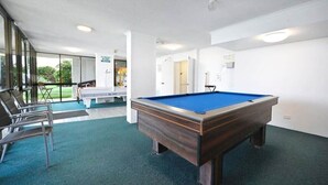 Uma mesa de bilhar em um ambiente com carpete azul, paredes brancas e uma porta que dá para uma varanda.