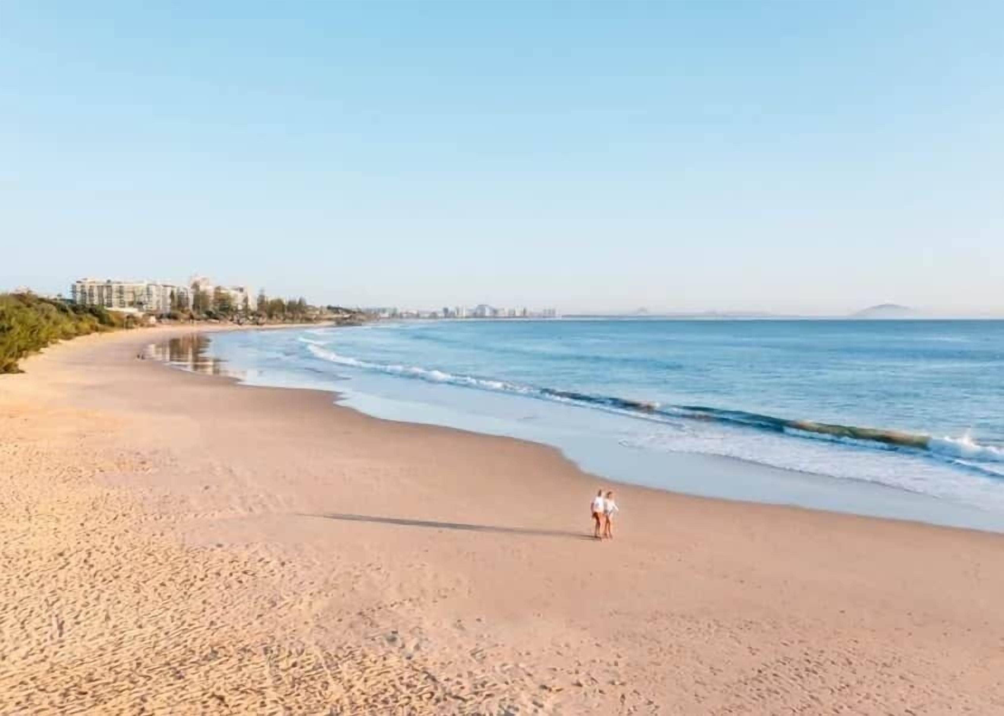 A beach with a person walking, clear skies, and buildings in the distance.