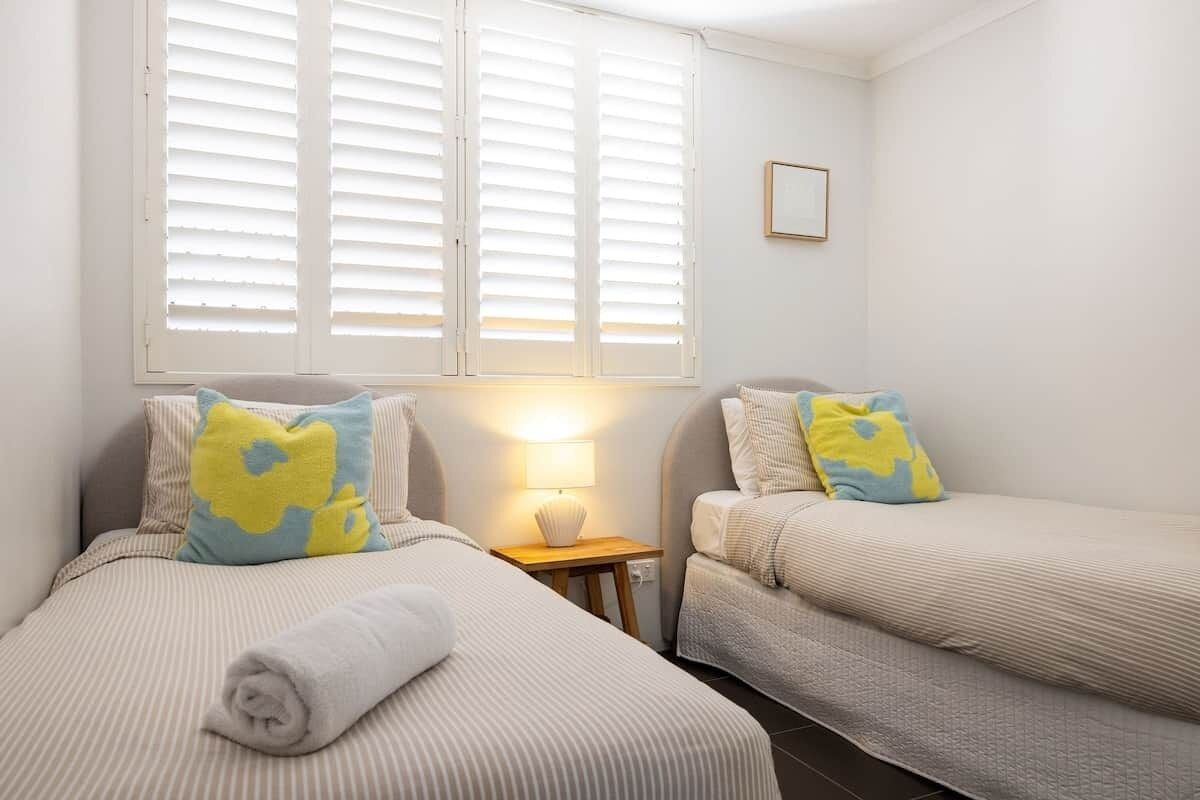 Une chambre avec deux lits, une table de chevet avec une lampe et une fenêtre avec des stores.