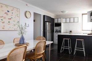 Eine moderne Küche mit Essbereich, ausgestattet mit einem weißen Tisch, Korbstühlen, einer gestreiften Vase und einem schwarzen Kühlschrank.
