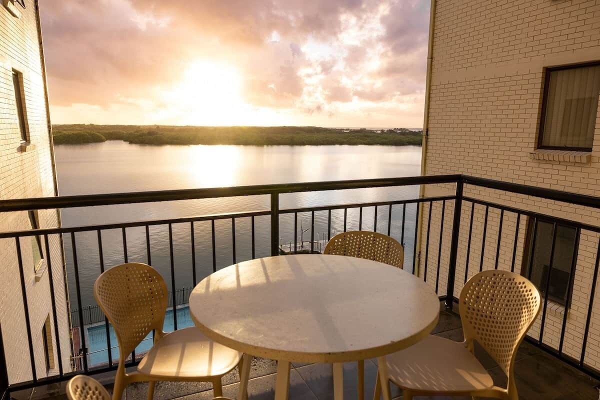 En balkon med et rundt bord og fire fletstole, med udsigt over et roligt vandareal og en skyet himmel.