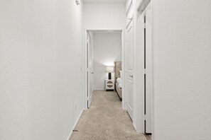 Un couloir étroit aux murs blancs et au sol recouvert de moquette, qui mène à une chambre à coucher équipée d’un lit et d’une table de chevet.