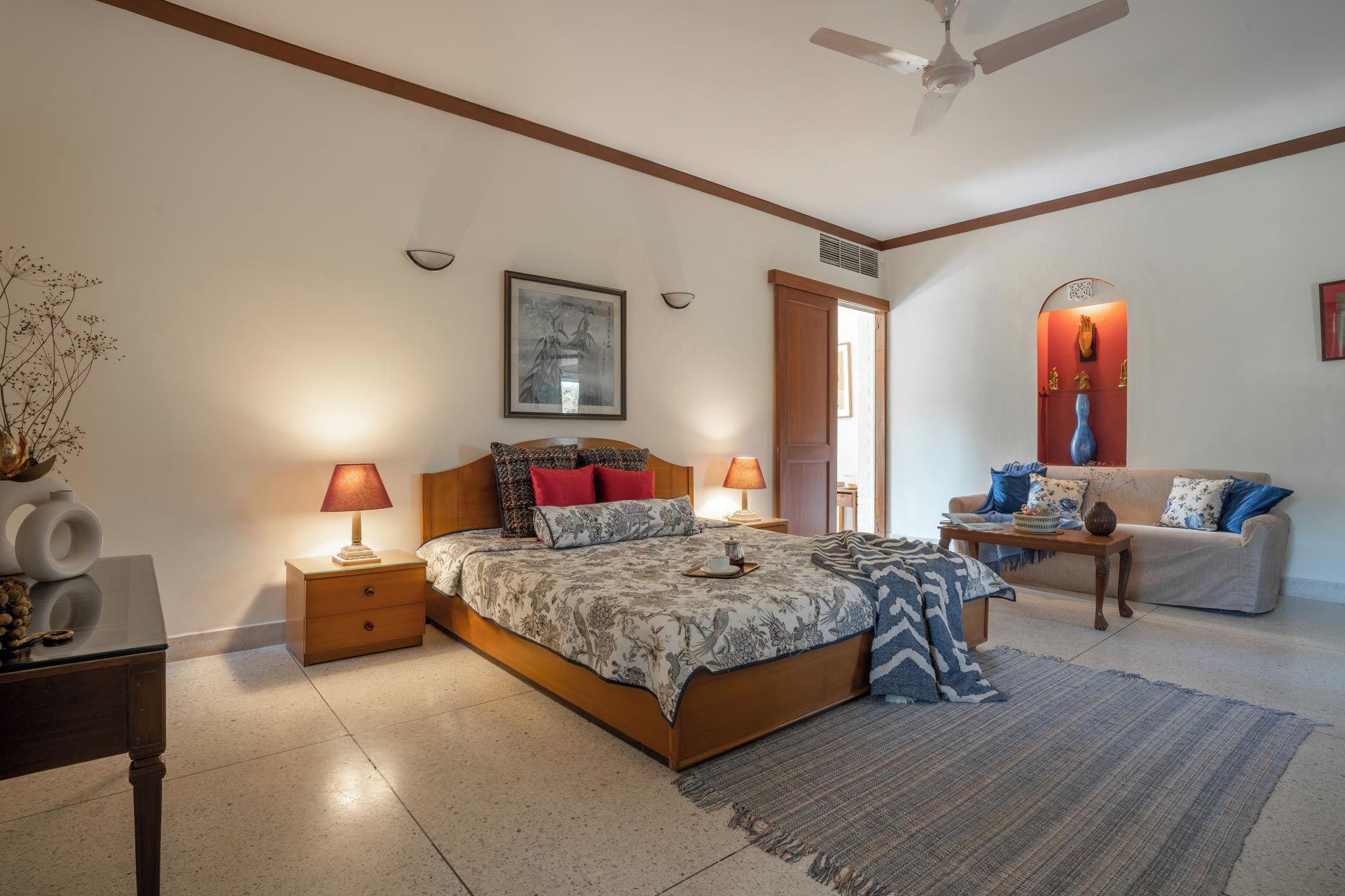 A bedroom with a bed, bedside tables, a nightstand, a chair, a sofa, a rug, a ceiling fan, and a wall-mounted shelf with decorative items.
