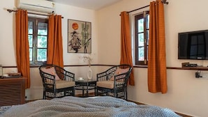 Un dormitorio con cama, televisión, ventana con cortinas, una silla y una mesa con un florero.