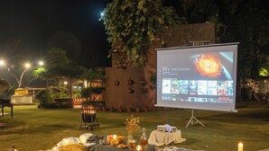 Montaje para una noche de cine al aire libre con una pantalla mostrando opciones de películas, mantas, almohadas y luces colgantes sobre un césped.