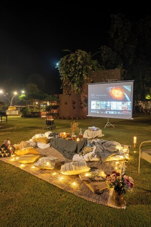 Montaje para una noche de cine al aire libre con una pantalla mostrando opciones de películas, mantas, almohadas y luces colgantes sobre un césped.
