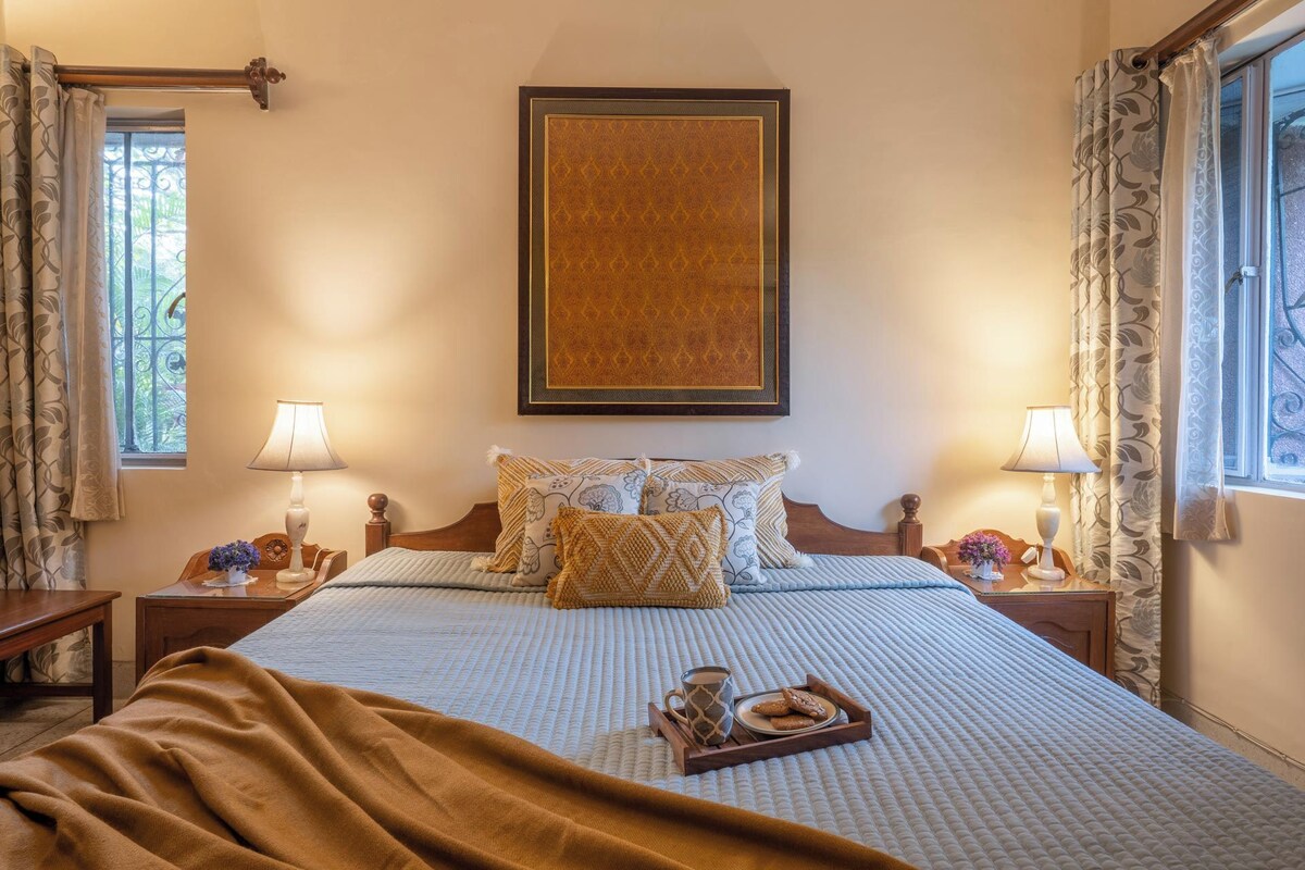 A neatly made bed with a tray of cookies and a mug, flanked by two bedside tables with lamps, a window with curtains, and a framed artwork above the bed.