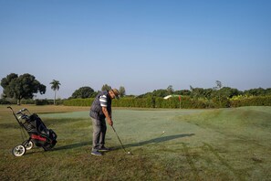 A golf course with a golfer preparing to take a shot, a golf bag, and a flag on the green. - The Homestead (Ramnagar)