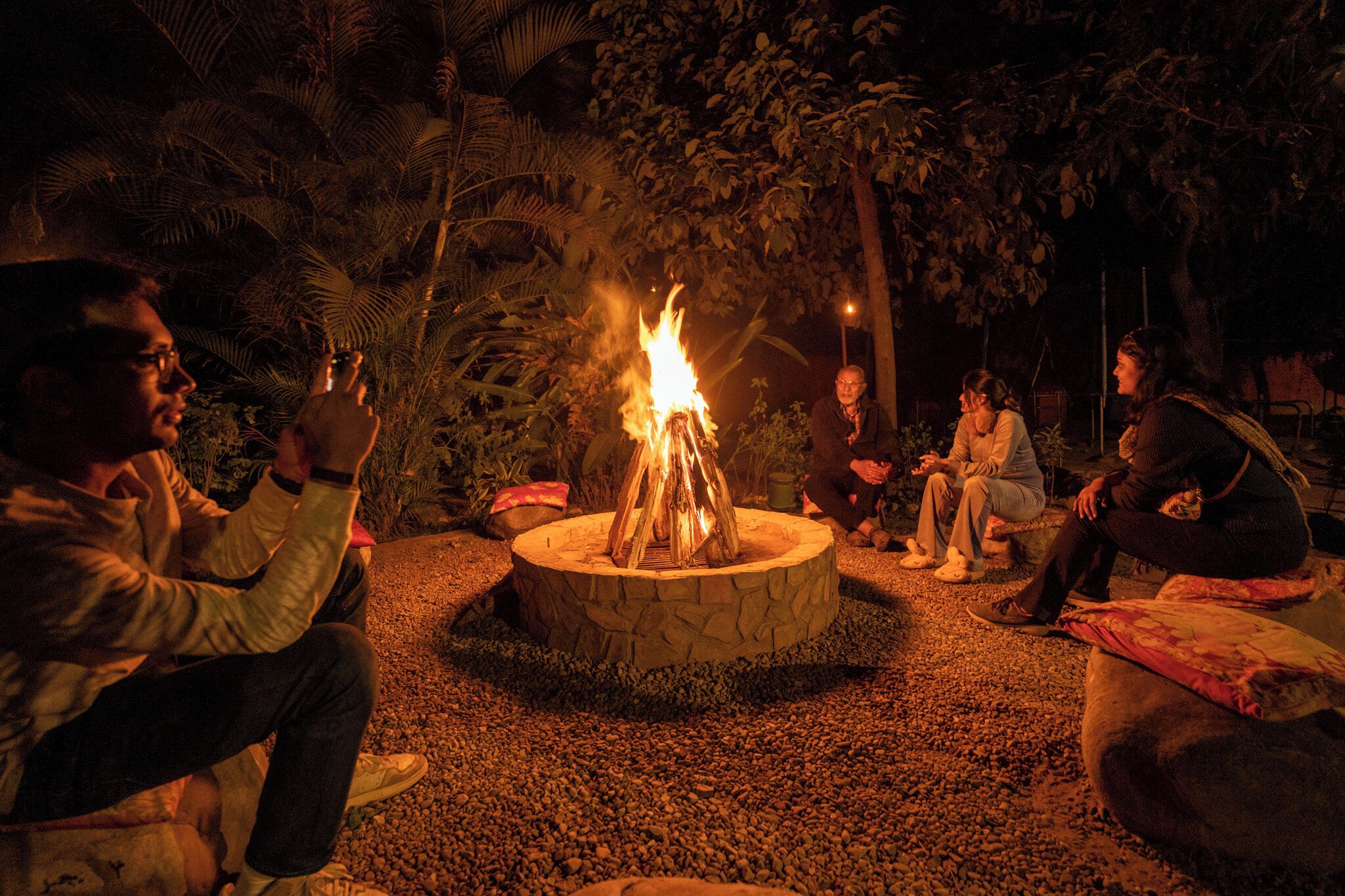 A group of people gathered around a fire pit in an outdoor setting with tropical vegetation.
