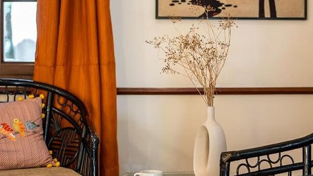 Una habitación con una cama, una mesa con una taza, un jarrón con flores secas y un cuadro en la pared.